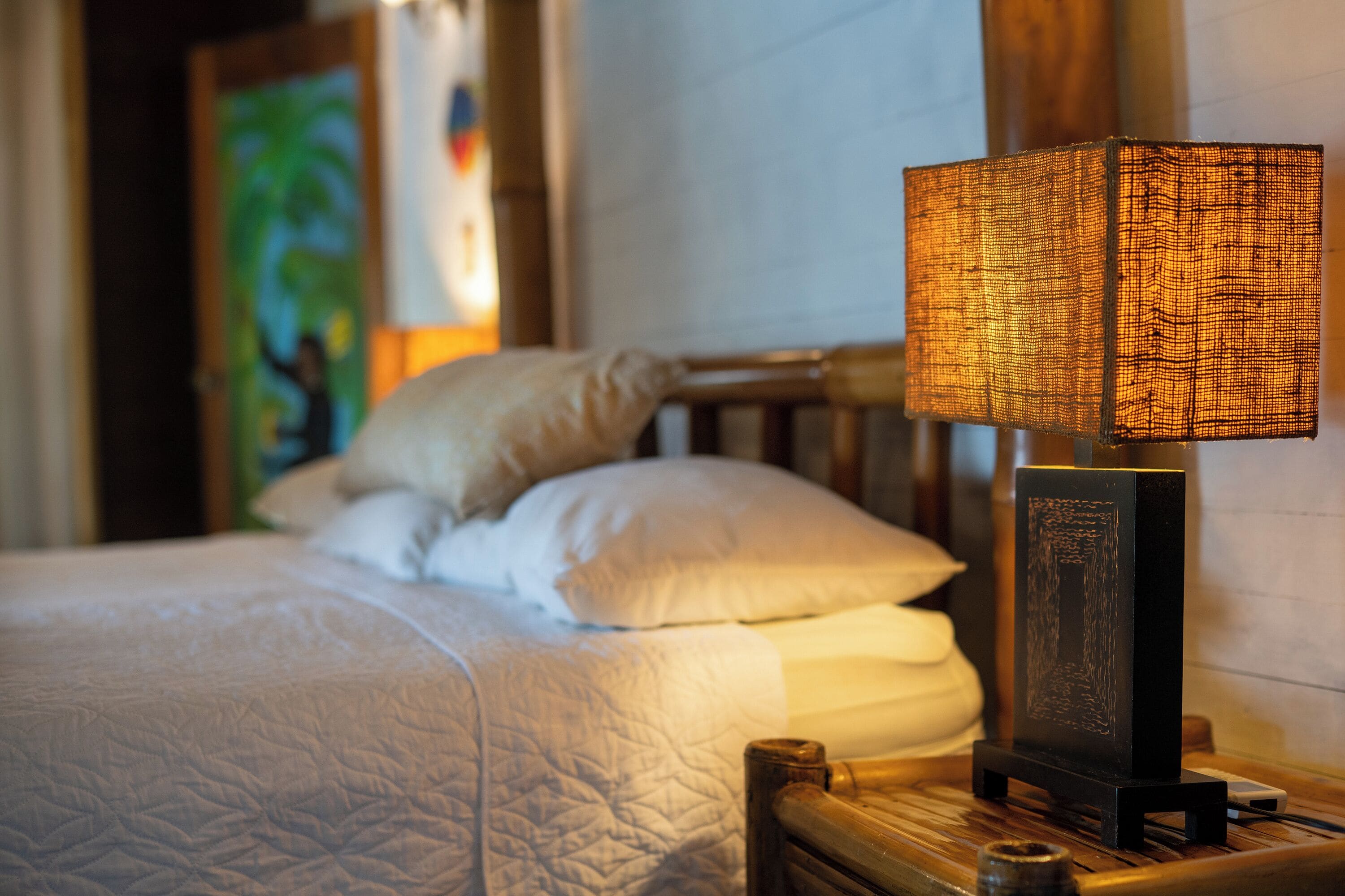 A neatly made bed with a white comforter, a wooden headboard, and a bedside lamp with a woven shade.