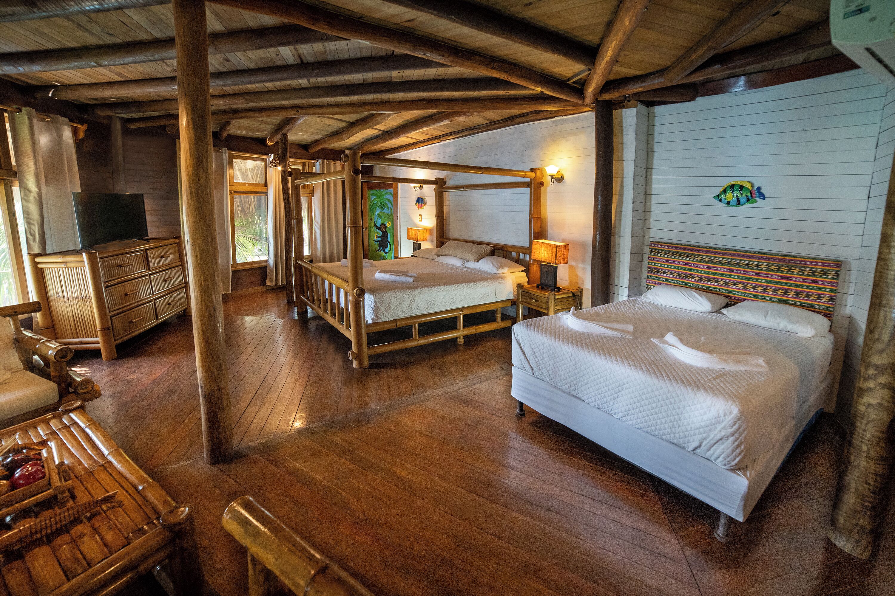 A wooden four-poster bed with white bedding, a wooden bench, and a television on a wooden dresser in a room with wooden floors and a sloped ceiling.