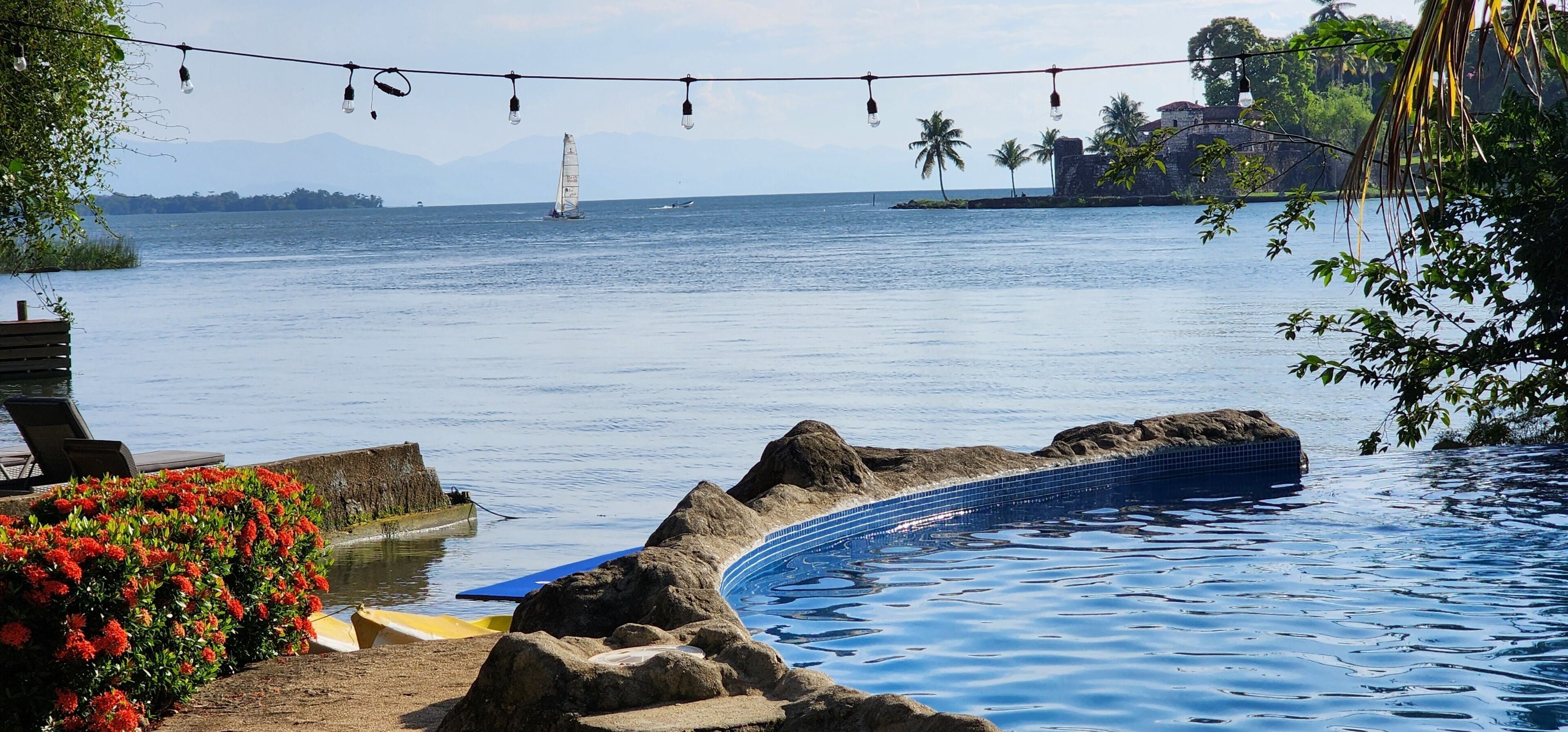 Una piscina de borde curvo, un velero en el agua y una cadena montañosa a lo lejos.