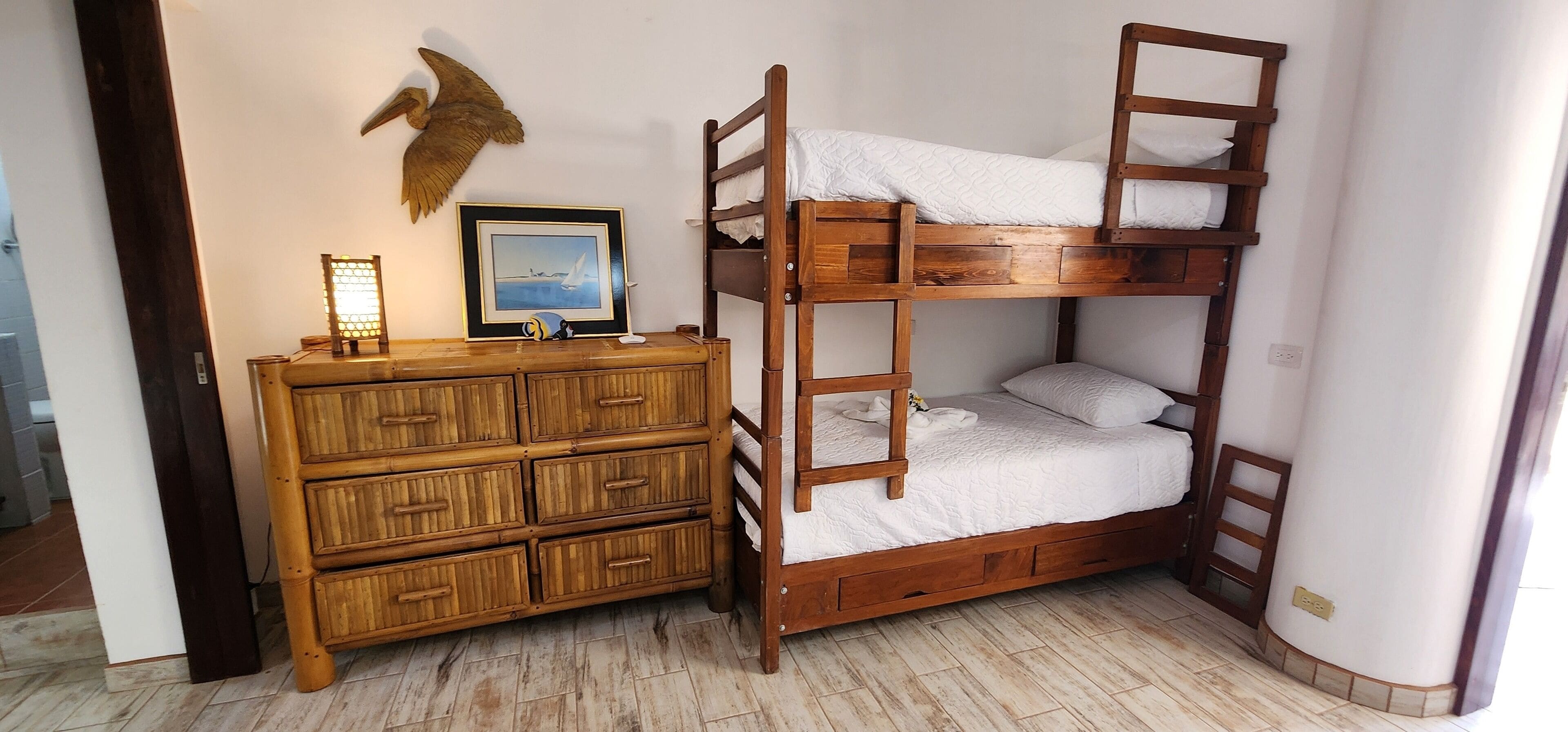 A bunk bed with a wooden ladder and drawers, a wooden dresser with multiple compartments, and a framed picture of a seascape on the wall.