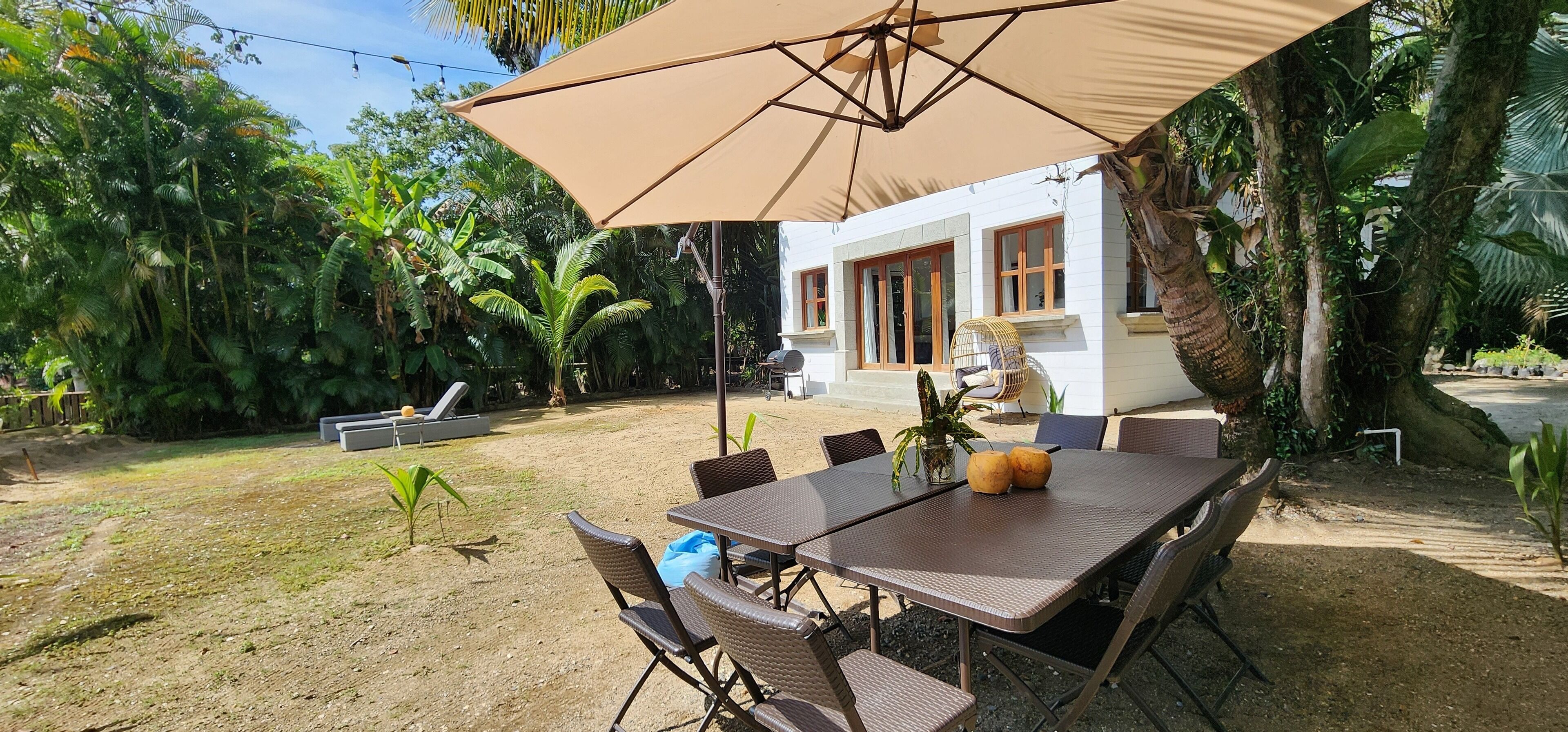 Un área de comedor al aire libre con una mesa y sillas bajo un paraguas grande, rodeada de exuberante follaje y un edificio blanco al fondo.