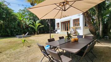 Un área de comedor al aire libre con una mesa y sillas bajo un paraguas grande, rodeada de exuberante follaje y un edificio blanco al fondo.