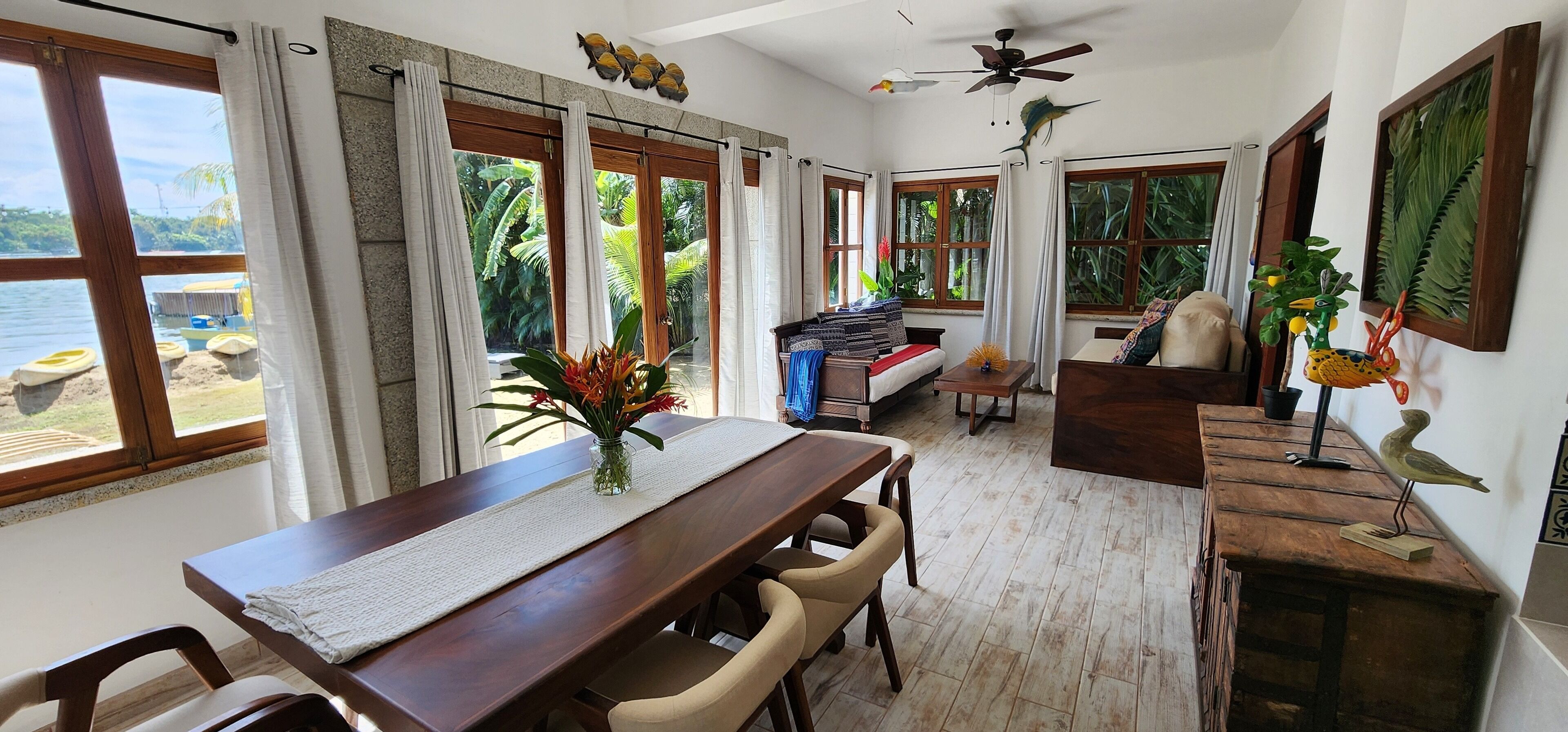 A spacious room with a wooden dining table, chairs, a sofa, and a ceiling fan. Large windows offer a view of the outdoors.