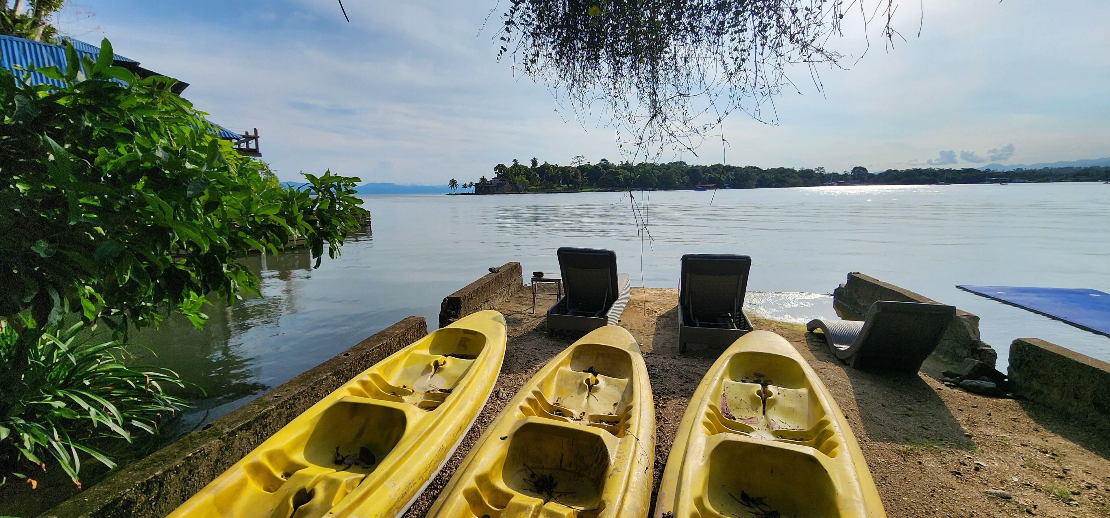 A calm lake with kayaks and chairs on a dock, surrounded by greenery and a distant shoreline.