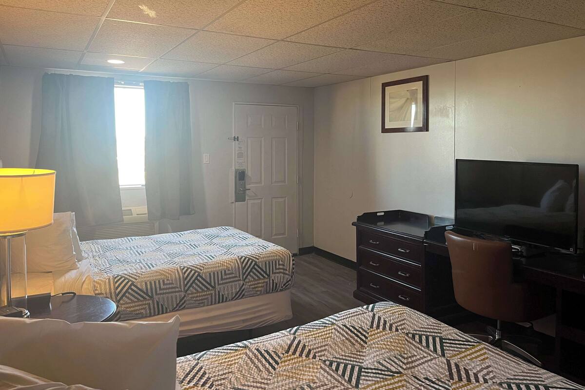 A hotel room with two beds, a desk with a television, a dresser, and a lamp.