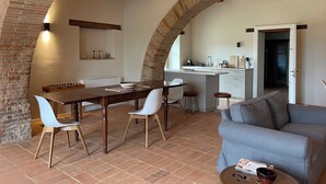 A dining area with a brick arch, wooden ceiling beams, a kitchen with a sink and cabinets, and a gray sofa. - Exclusive farmhouse with heated swimming pool (Marsciano)