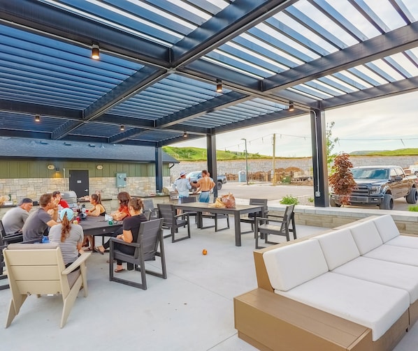 Un patio exterior con mesas y sillas, una zona de parrilla y un área de estar cubierta con vista al estacionamiento.