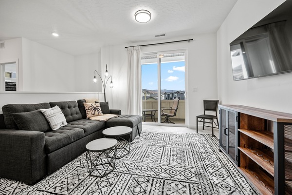 The living room has plush seating, an expansive smart TV, and sliding patio doors that lead to the enclosed balcony.