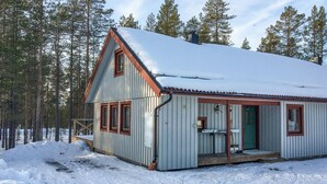 A small, single-story cabin with a steeply pitched roof, surrounded by a snowy landscape and tall pine trees. - Amazing apartment in Idre with sauna (Idre)