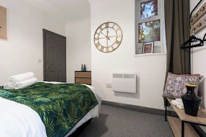 A hotel room with a bed, a wall clock, a window, a heater, and a small table with a vase.