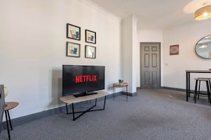 A modern hotel room with a flat-screen TV on a wooden stand, a small side table, and a dining area with a table and chairs. - Sienna's Apartment -2 Bed getaway in Pontypool (Pontypool)