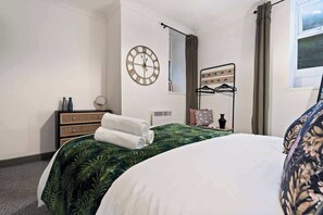 A hotel room with a bed, a wall clock, a mirror, a window with curtains, and a bedside table with a lamp and a vase.
