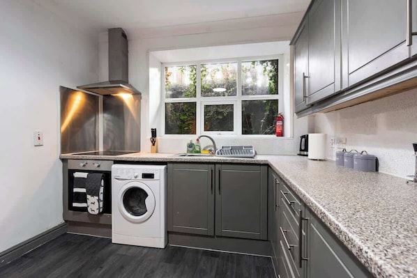 A compact kitchen with a built-in washer, a window with a view of greenery, and a stainless steel range hood.