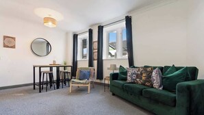 Ett vardagsrum med en grön soffa, en rund spegel, ett litet bord med pallar och ett fönster med gardiner.