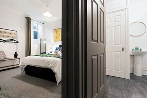 A hotel room with a bed, a bedside table, a mirror, a window with curtains, and a door leading to a bathroom with a sink and toilet.