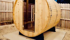Una sauna de madera con puerta de vidrio, sobre base de hormigón, rodeada por una valla de madera.