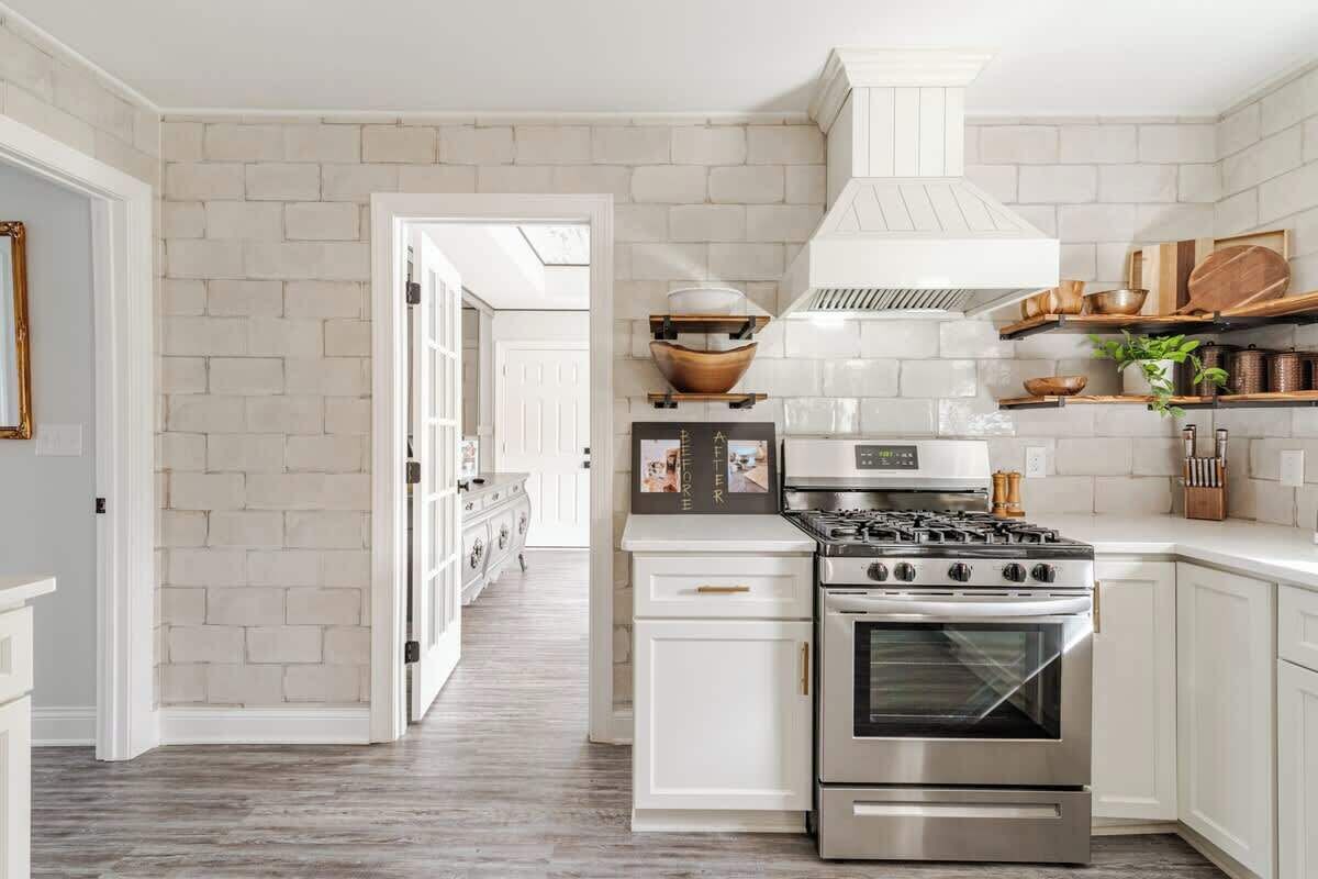 Una cocina con gabinetes blancos, estufa de acero inoxidable y salpicadero de ladrillo blanco.