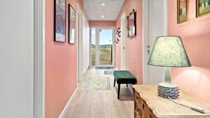 Un pasillo con paredes rosadas, cuadros enmarcados, un banco verde y una mesa con lámpara.