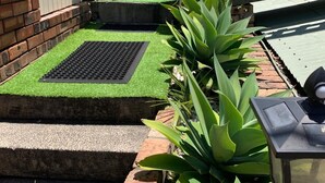 A backyard with a white fence, a brick wall, and a small garden area with green plants and a metal grate. - Gorgeous Apartment in Kiama, minutes from Kiama's famous Blowhole & Beaches (Kiama)