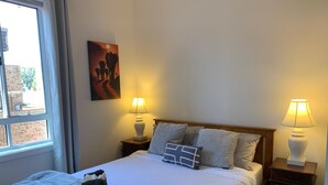 A neatly made bed with a quilt, pillows, and a blanket. There are two bedside tables with lamps. A painting hangs on the wall above the bed. - Gorgeous Apartment in Kiama, minutes from Kiama's famous Blowhole & Beaches (Kiama)