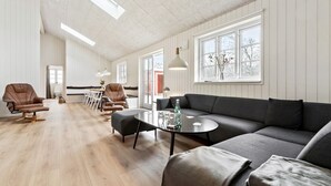 A spacious living area with a wooden floor, a grey sofa, two leather armchairs, a dining table with chairs, and large windows providing natural light.