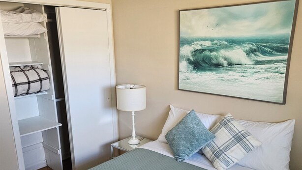 A hotel room with a bed, a painting of waves, a closet, and a nightstand with a lamp.