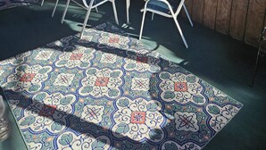 A covered patio with a table and chairs, a patterned rug, and a view of trees through large windows.