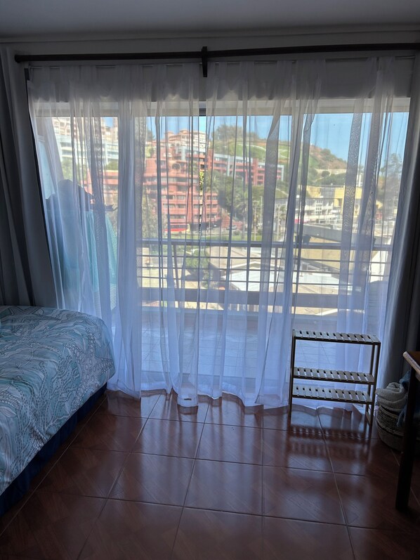 Un dormitorio con una cama, una estantería de madera y vistas a edificios a través de cortinas traslúcidas.