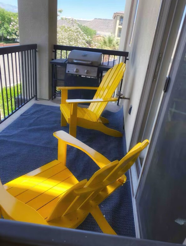 A balcony with a blue mat, two yellow Adirondack chairs, and a grill.