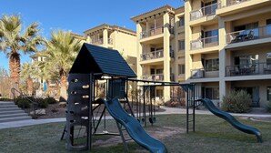 Un parc infantil amb tobogan i cadira de balanceig, envoltat d'una zona enjardinada i palmeres, i un edifici de diversos pisos al fons.