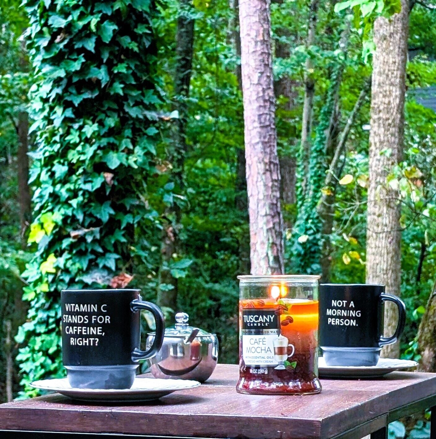 Une table avec trois objets : une tasse noire avec du texte, une bougie en verre étiquetée « Bougie de Toscane » et une tasse noire avec du texte.