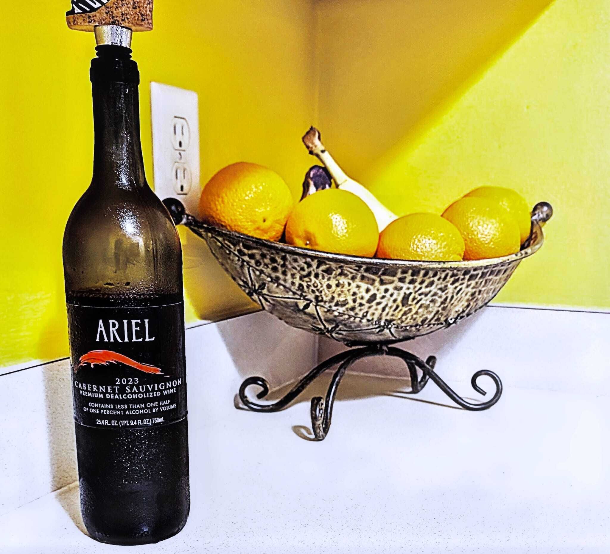 Une bouteille de vin Ariel 2023, un Cabernet Sauvignon, est posée à côté d’un bol en métal contenant des citrons et des oranges, sur une table blanche, le tout sur fond d’un mur jaune.
