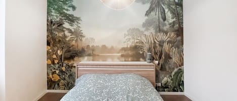 A bedroom with a bed, a wall mural of a tropical landscape, and a hanging light fixture.