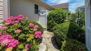 Un chemin menant à une maison dotée d’un porche abrité, entourée d’une végétation luxuriante et de plantes en fleurs.