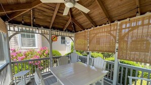 Une véranda abritée avec une table et des chaises, un ventilateur de plafond et une vue sur une maison ornée de fleurs roses.