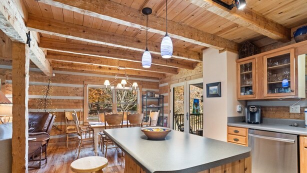 A rustic cabin interior with a wooden dining area, a kitchen with wooden cabinets, and a large window offering a view of the outdoors. - Charming Cabin in Seven Devils (Seven Devils)