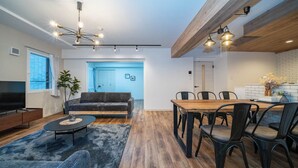 Ein modernes Wohnzimmer mit Sofa, Couchtisch und Fernseher, ein Essbereich mit Holztisch und Stühlen sowie eine Küche mit weißer Arbeitsplatte und Blumen.