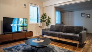 Ein modernes Wohnzimmer mit Flachbildfernseher, grauem Sofa, Couchtisch aus Holz und großem Fenster mit Vorhängen.