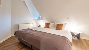 Quarto moderno com teto inclinado, cama grande com cobertor marrom, almofadas brancas e uma pequena mesa de cabeceira com abajur.
