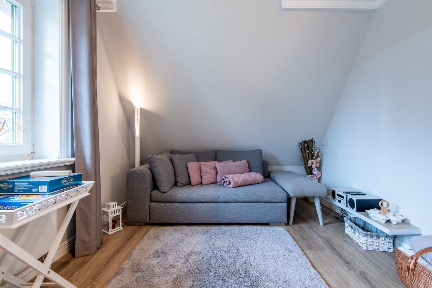 Une pièce chaleureuse avec un canapé gris, une petite table avec des livres, une lampe et une fenêtre avec des rideaux.