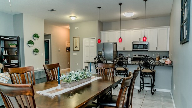 Un comedor con mesa y sillas de madera, una cocina con armarios blancos y electrodomésticos de acero inoxidable, e iluminación colgante.