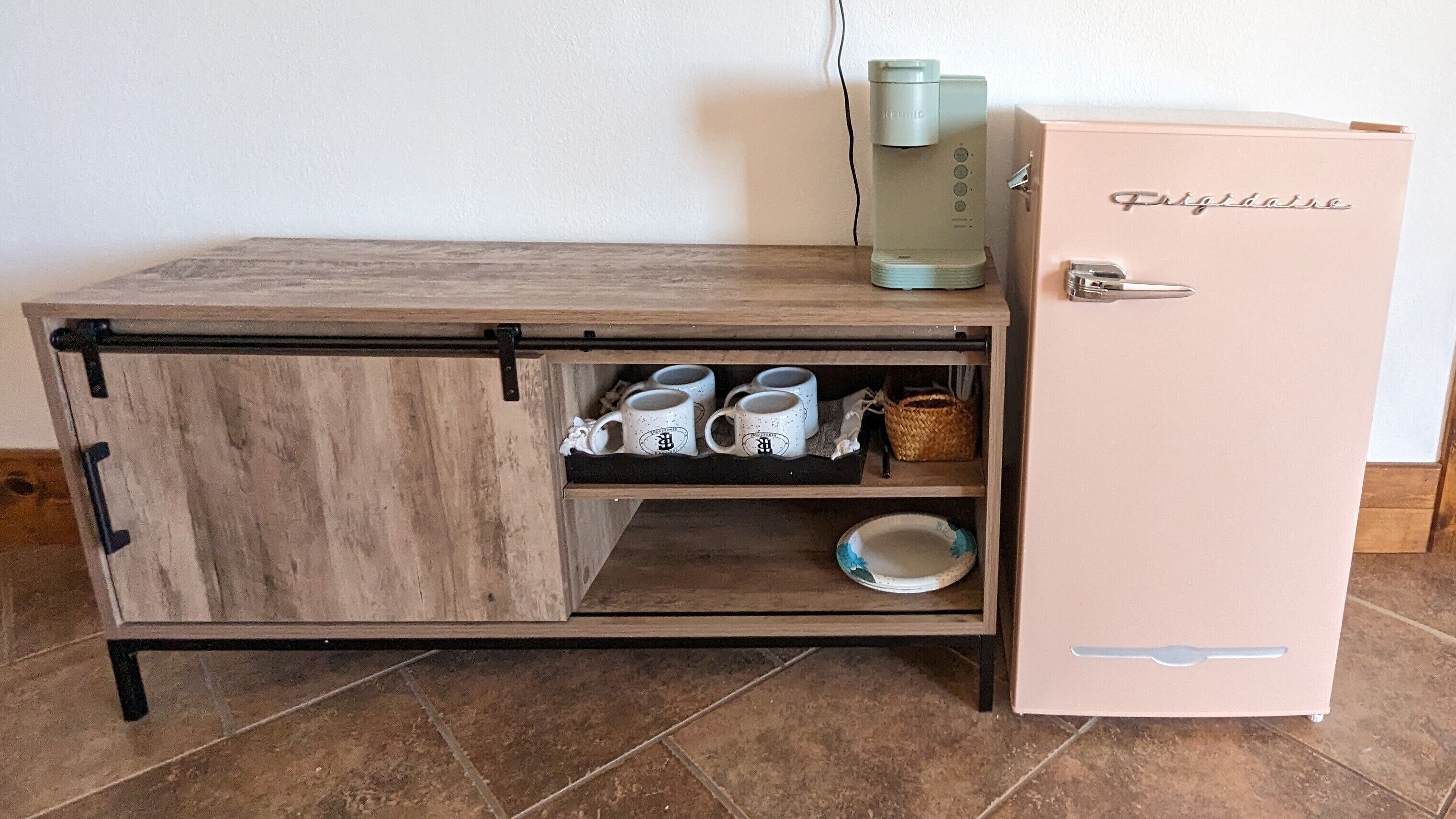 A kitchenette with a wooden countertop, a coffee maker, a refrigerator, and a shelf with mugs.