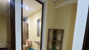 Un corridoio con un bagno visibile attraverso una porta aperta. Il bagno è dotato di toilette, lavandino e zona doccia. Sul pavimento c'è un secchio blu e uno scaffale in legno con piante in vaso.