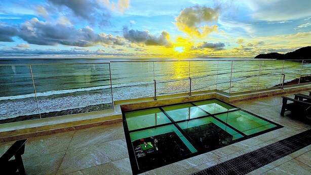 Área externa ensolarada com piso de vidro que dá para uma piscina, cercada por corrimão e bancos.