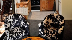 Et kompakt køkken med træskabe, en mikroovn, komfur og et vindue med udsigt til grønne omgivelser. I opholdsområdet er der to stole med leopardprint og et lille rundt bord.
