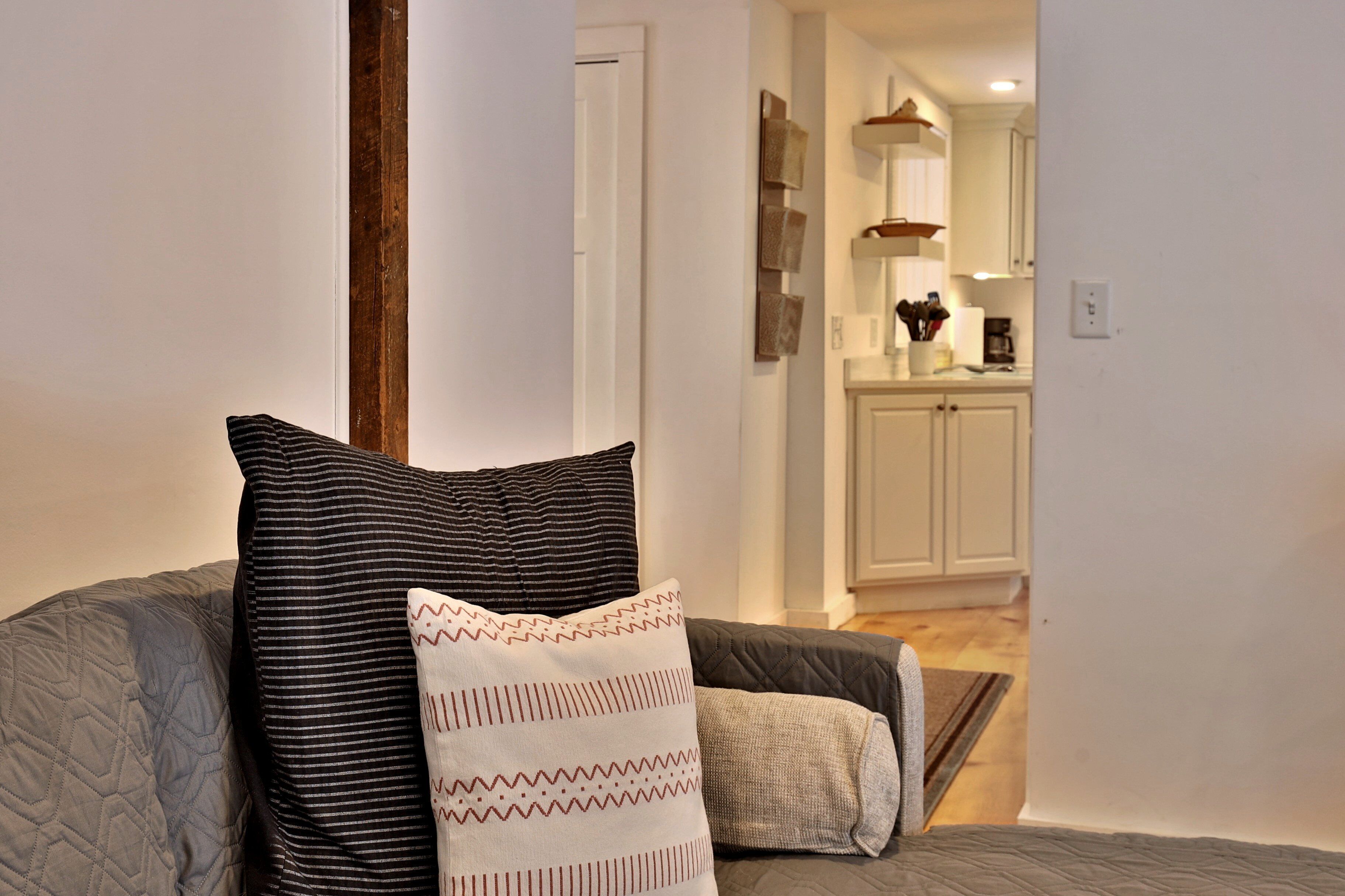 A living room with a grey couch, two pillows, and a view into the kitchen area.