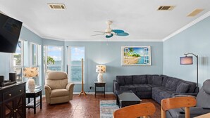 A living room with a large window, a ceiling fan, a dark-colored sofa, a beige armchair, and a television on a stand. - Sailfish Haven #390 - Ocean Views, 35' seawall (Cudjoe Key)