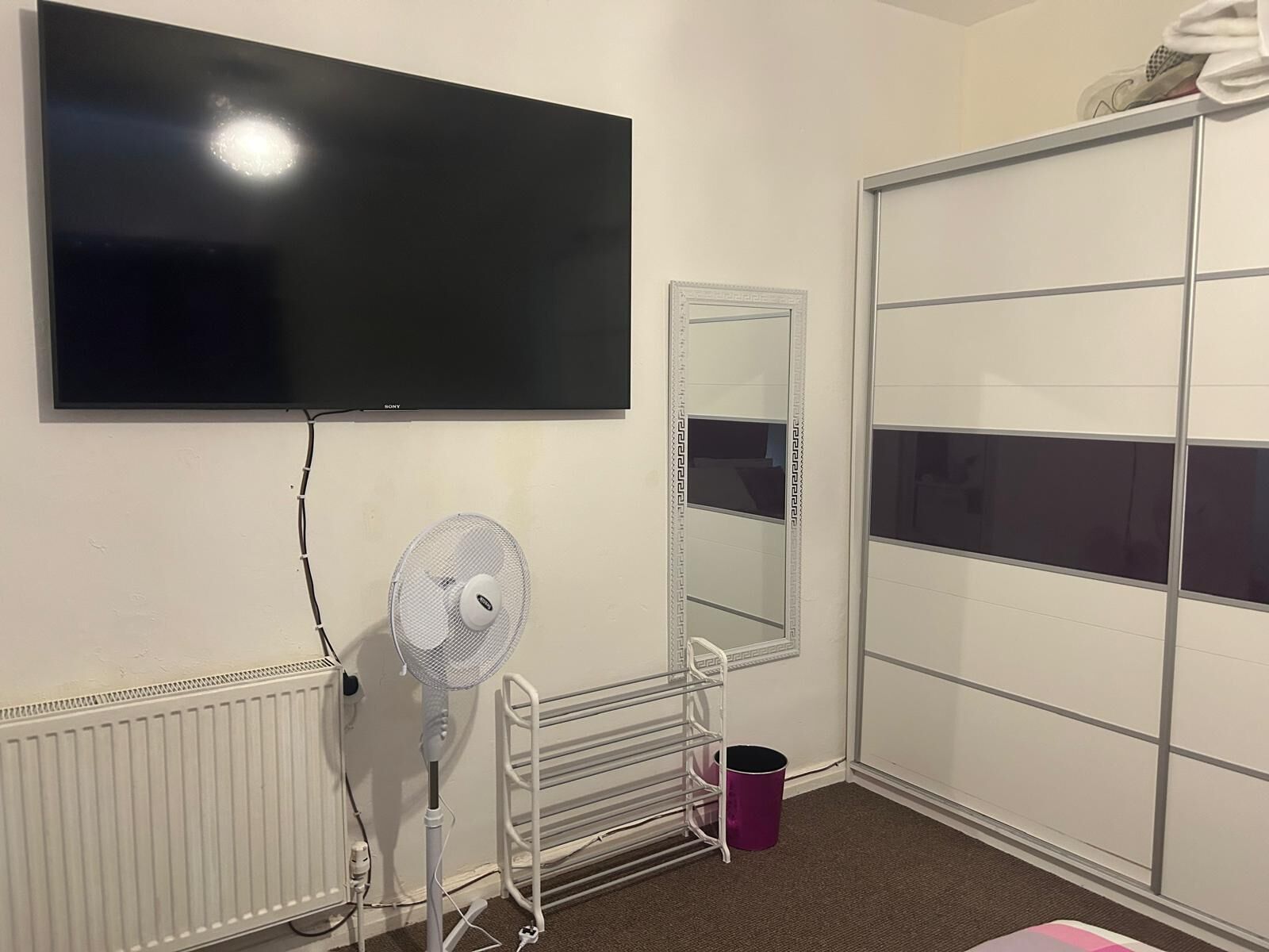A room with a large flat-screen TV mounted on the wall, a standing fan, a radiator, a mirror, and a sliding closet door.