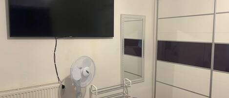 A room with a large flat-screen TV mounted on the wall, a standing fan, a radiator, a mirror, and a sliding closet door.