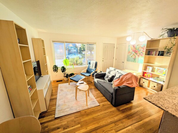A living room with a sofa, a coffee table, a bookshelf, and a window with a view of the street. - Bright 2BR, Cal King Bed, 15 Min walk to OSU (Corvallis)